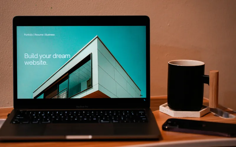 A laptop on a wooden desk showing a website homepage, next to a black mug and a phone.