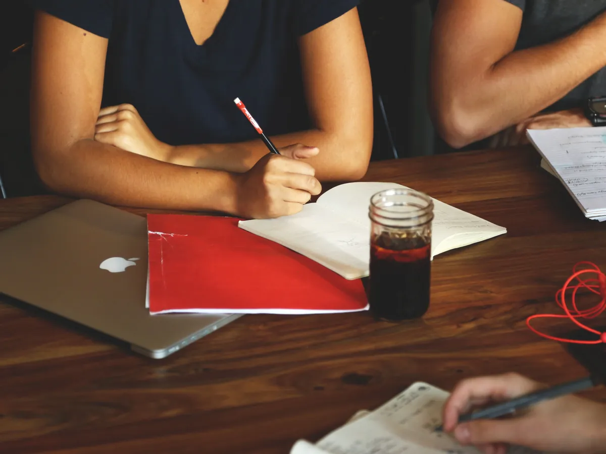 Three people at a wooden table taking notes, with notebooks, a red folder, a closed laptop and a glass of iced coffee.
